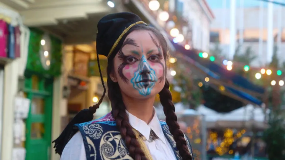 Person in Tracht mit bunter Gesichtsbemalung vor festlicher Kulisse.