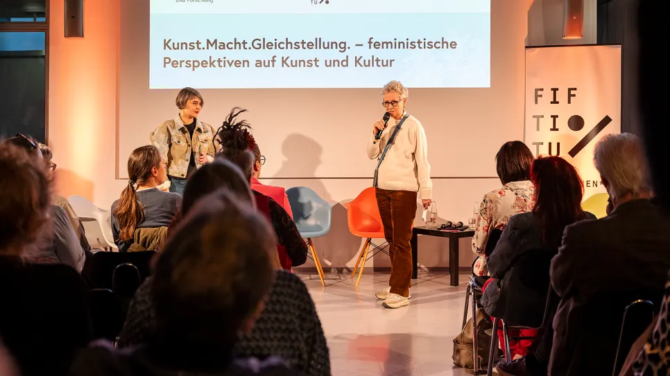 Zwei Frauen auf Bühne, halten Vortrag vor Publikum, feministische Themen, beleuchteter Raum.