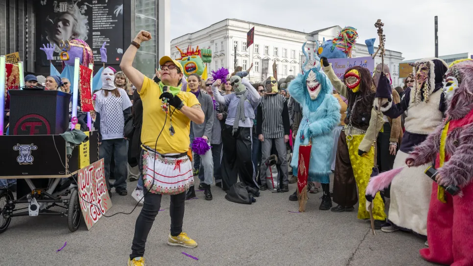 Protest mit bunten Kostümen und Masken, Person in gelbem Shirt hält Mikrofon.