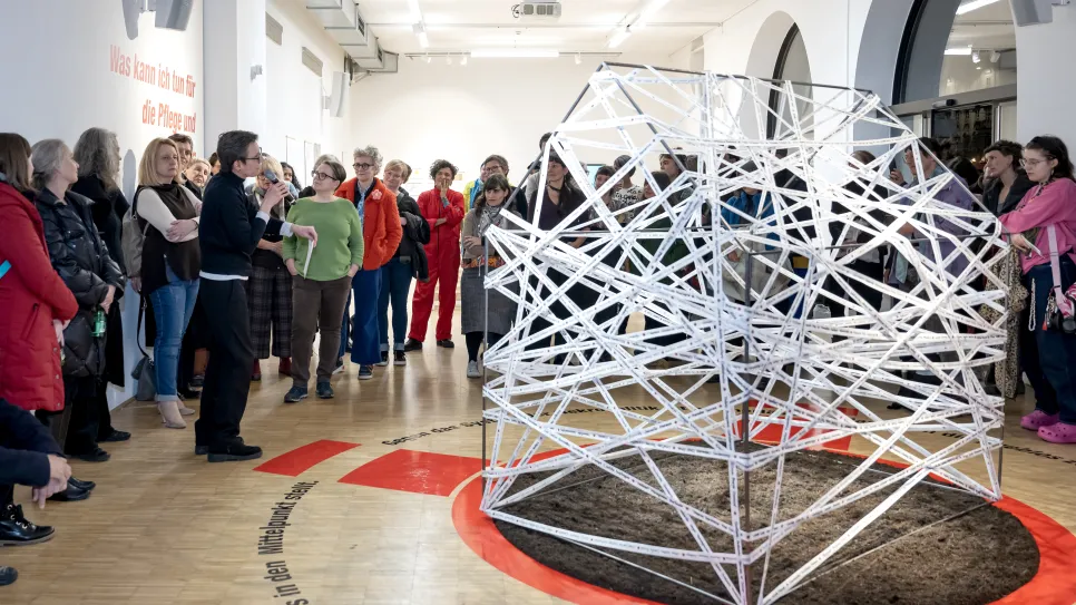 Große Gruppe Menschen betrachtet eine komplexe, künstlerische Installation in einem hellen Raum.