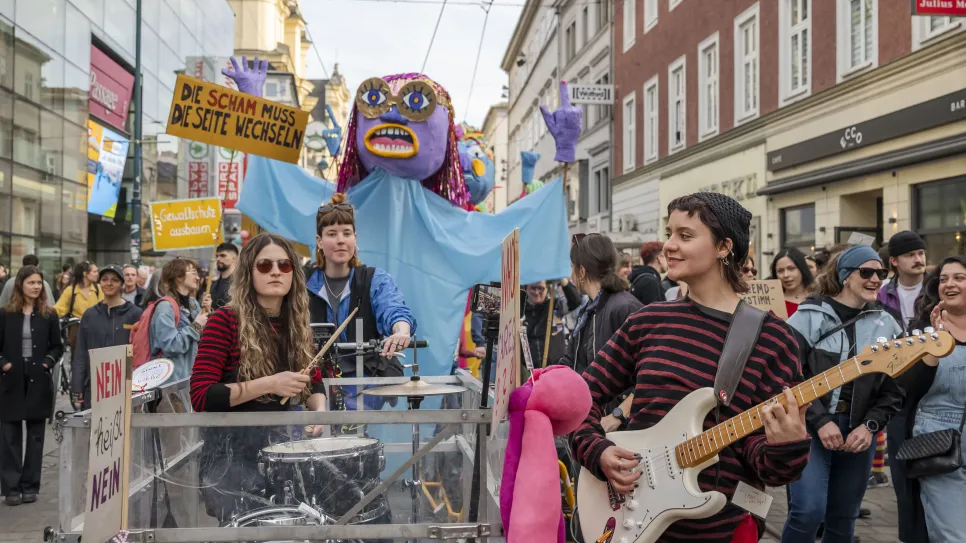 Menschen bei einer feministischen Demo, mit Musik und farbenfrohen Schildern in einer Stadtstraße.