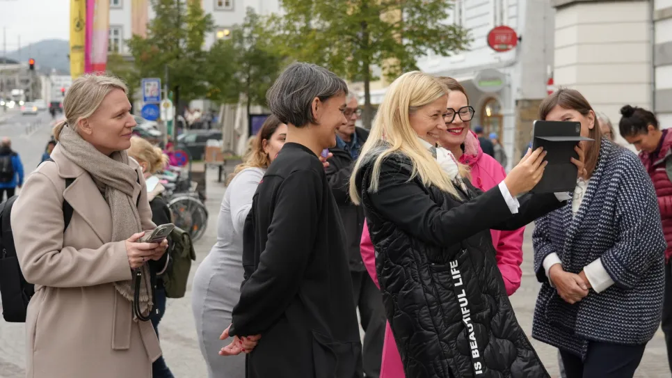 Frauen in einer Gruppe, eine hält ein Tablet, andere lächeln; Stadtumgebung im Hintergrund.