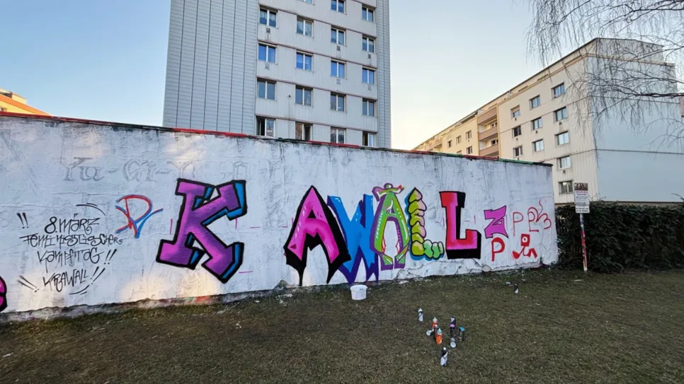 Graffiti "KAWAL" auf Betonwand vor Hochhäusern, bunte Buchstaben, sonniger Tag.