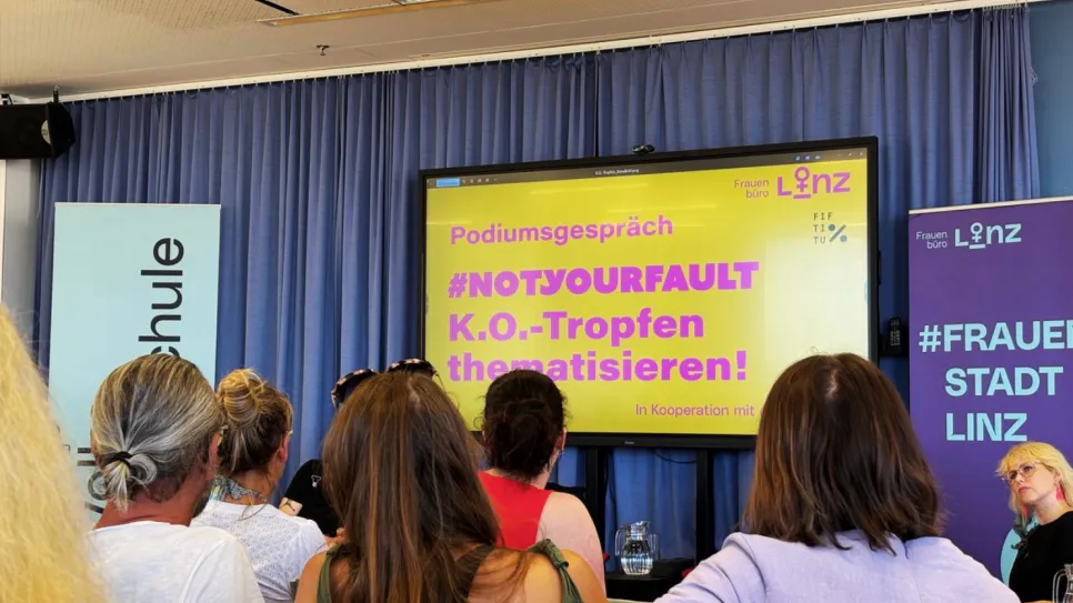 Podiumsgespräch mit Publikum, Thema: K.O.-Tropfen, gelber Bildschirm, blaue Vorhänge.