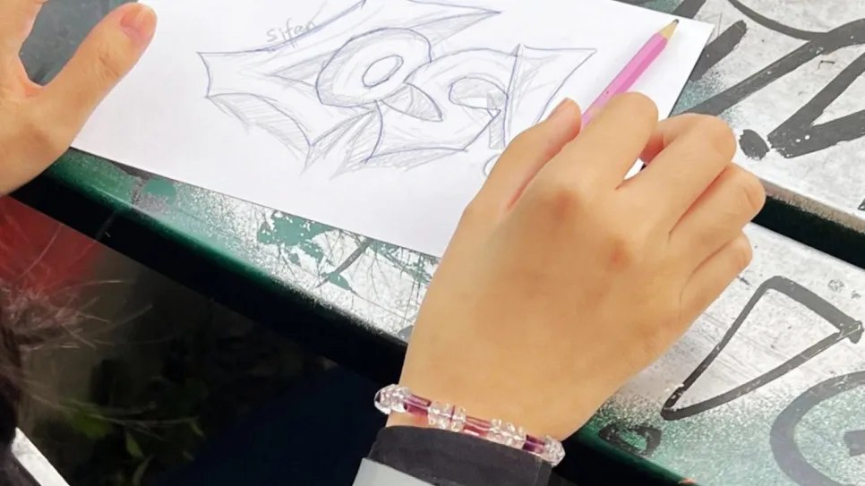 Hände zeichnen mit Bleistift auf Papier, Grafitti-Tisch im Hintergrund.