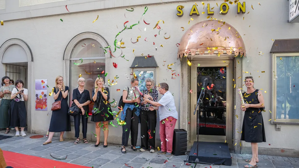 Menschen feiern vor einem Gebäude mit der Aufschrift "Salon", Konfetti in der Luft.