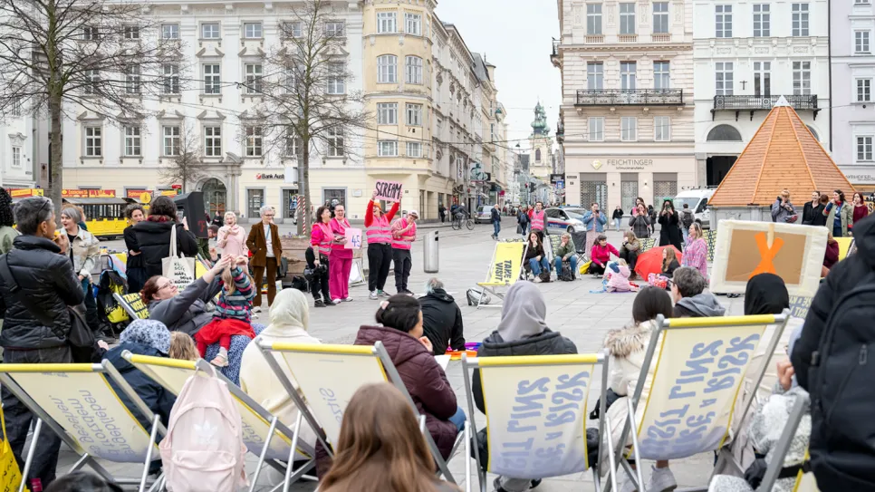 Versammlung von Menschen auf einem Stadtplatz mit Liegestühlen und Plakaten.