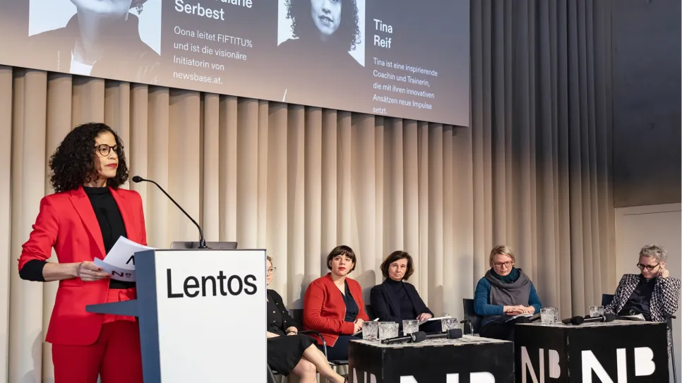 Rednerin im roten Anzug am Podium, fünf Frauen sitzen, minimalistischer Raum.