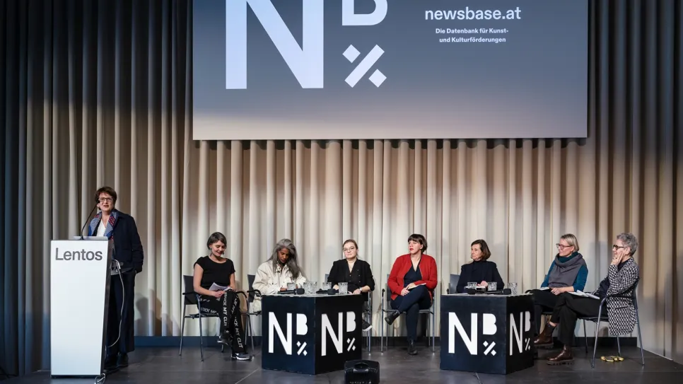 Podiumsdiskussion mit sieben Frauen auf einer Bühne, Logo "NB" im Hintergrund.