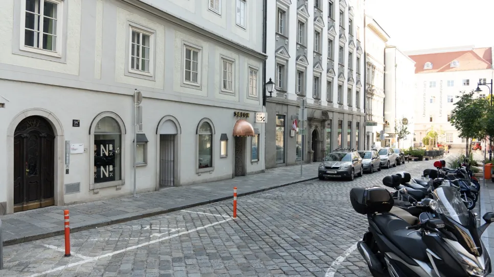 Pflasterstraße mit geparkten Autos und Rollern, Häuserfassaden rechts.