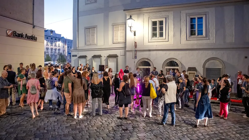 Menschenansammlung auf gepflasterter Straße bei Dämmerung, diskutierend vor beleuchtetem Gebäude.