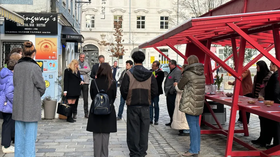 Menschenmenge bei einem roten Stand auf Kopfsteinpflasterstraße, städtische Umgebung.