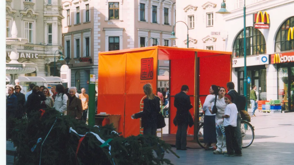 Rote Informationshütte auf belebtem Stadtplatz. Menschen stehen davor und unterhalten sich.