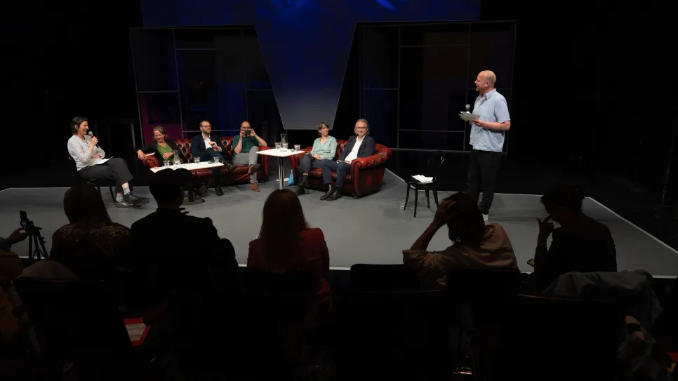 Diskussionsrunde mit sieben Personen, eine spricht stehend; Theaterbühne, abgedunkelt.