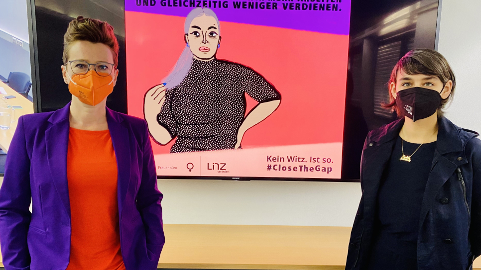 Zwei Frauen mit Masken vor feministischem Plakat zur Gender Pay Gap.