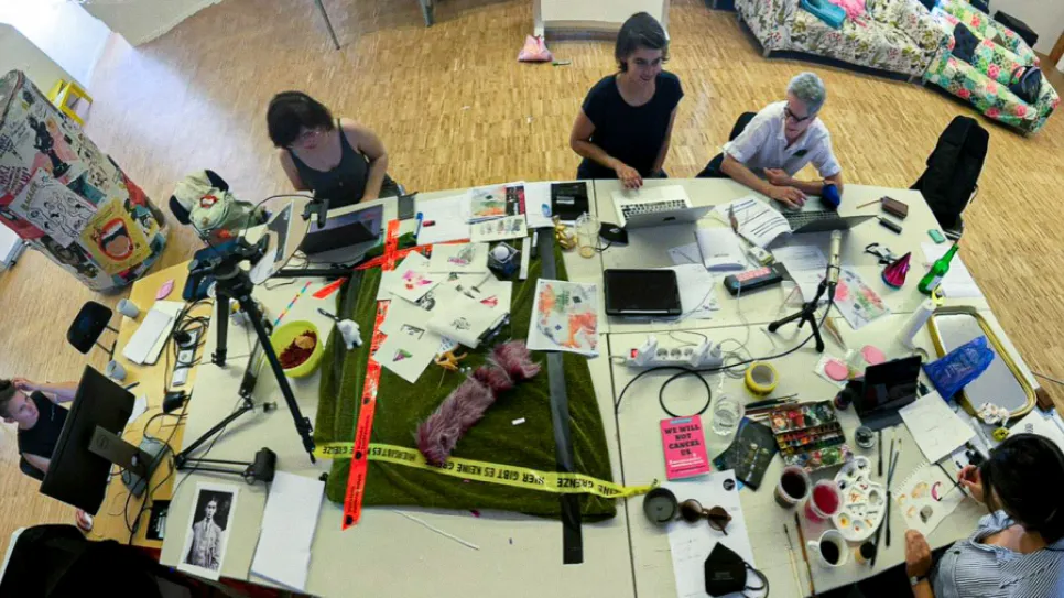 Fünf Frauen arbeiten kreativ an einem großen Tisch voller Materialien und Laptops.