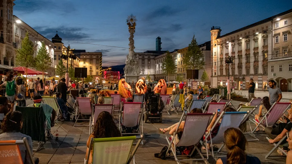 Menschen sitzen auf Liegestühlen bei Abenddämmerung auf einem belebten Platz.