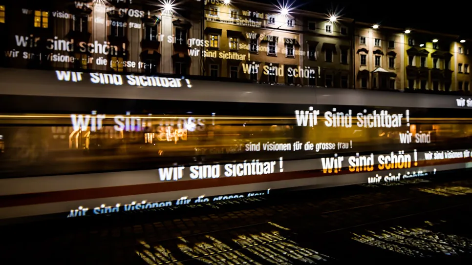 Lichter mit "Wir sind sichtbar!" auf vorbeifahrender Tram bei Nacht.