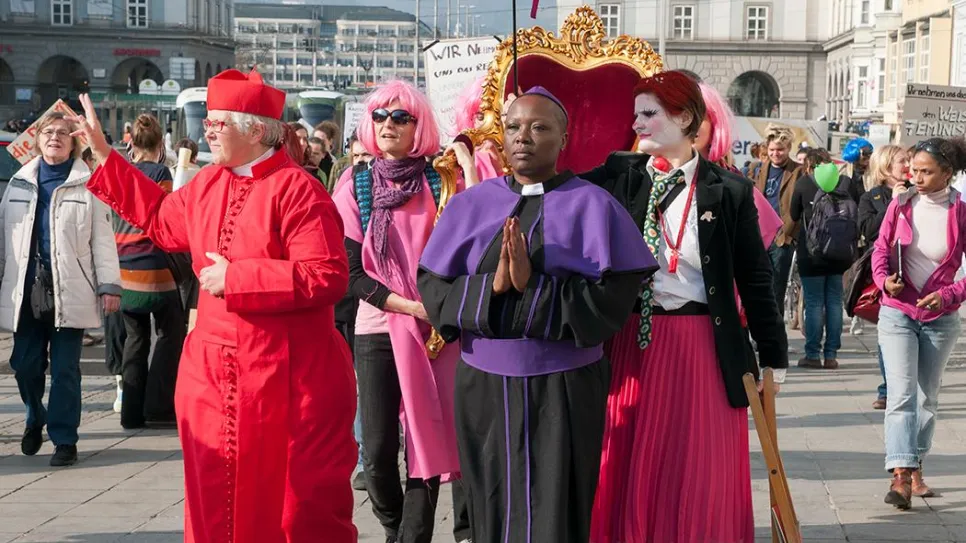 Menschen in bunten Outfits bei einer Straßenparade mit pinkem Schirm und Thron.