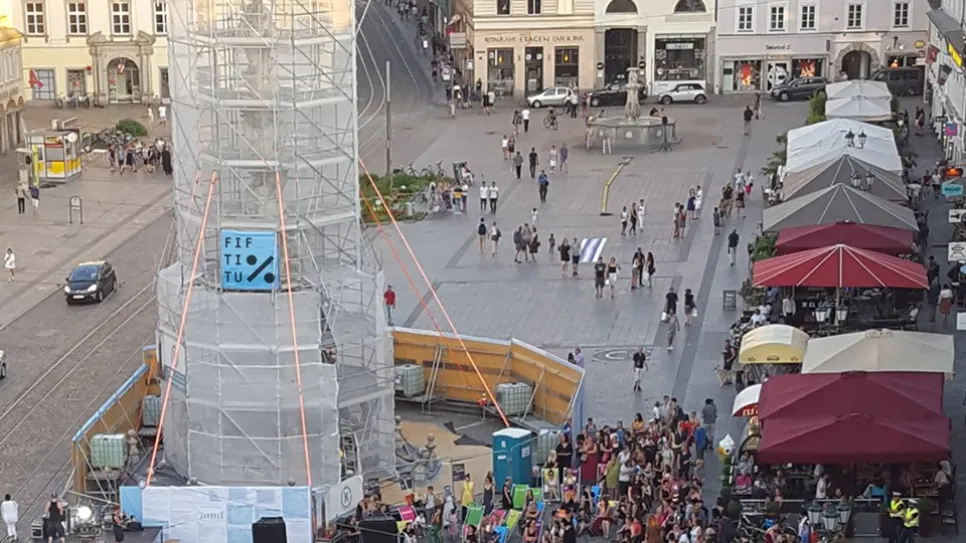 Menschenmenge um eine verhüllte Statue, bunte Schirme, helle Gebäude im Hintergrund.