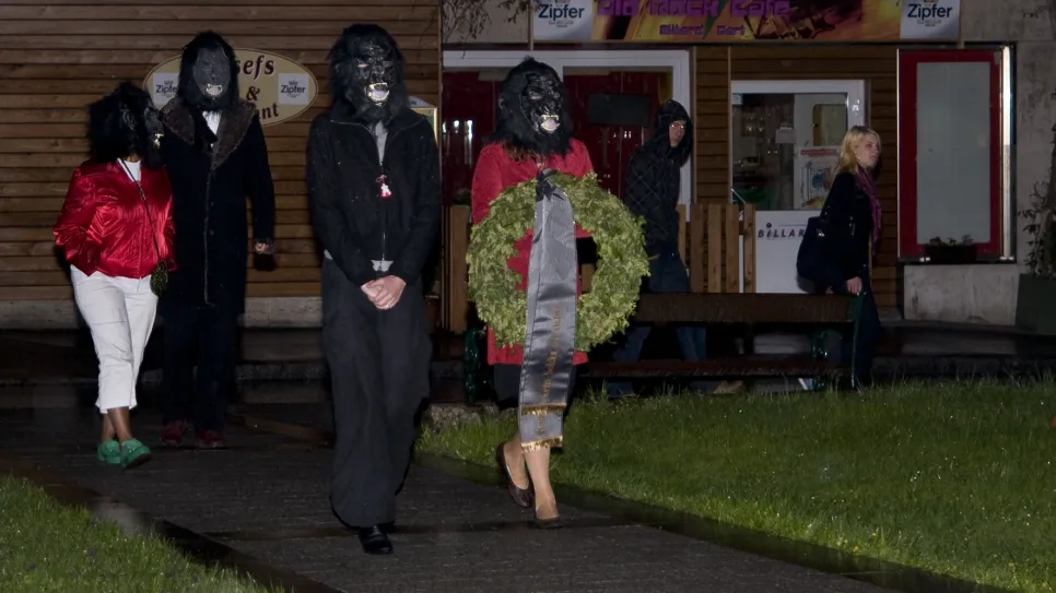 Menschen in Kostümen mit Masken und einem Kranz gehen einen Gehweg entlang, Nachtaufnahme.