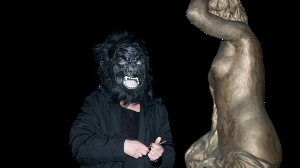 Person mit Gorilla-Maske neben goldener Statue bei Nacht.