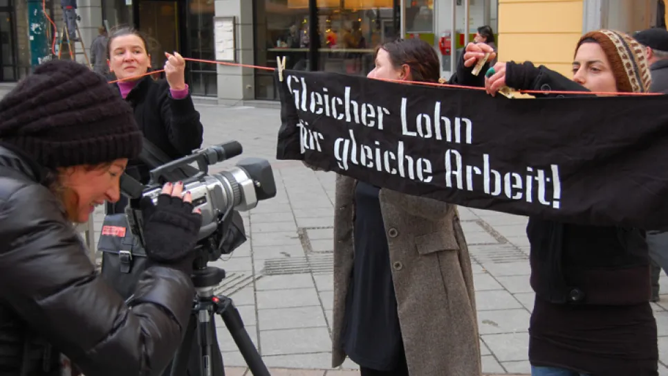 Personen halten Banner mit Aufschrift, eine filmt mit Kamera, städtische Kulisse.