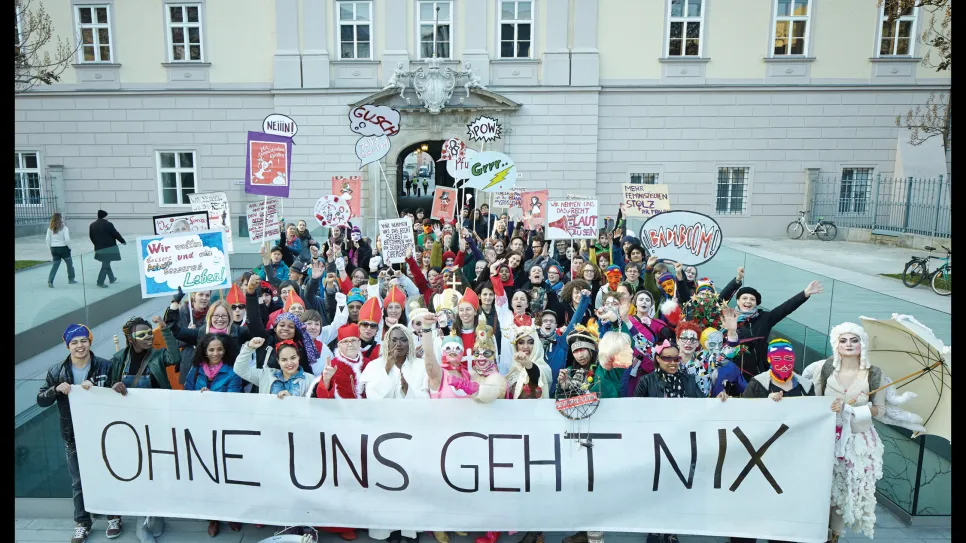 Große Menschengruppe mit Protestplakaten und Banner vor einem Gebäude.