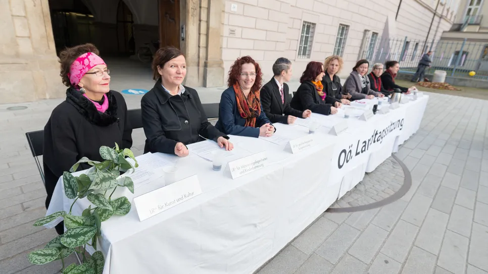 Neun Frauen sitzen an einem langen Tisch mit weißem Tuch vor einem Gebäude.