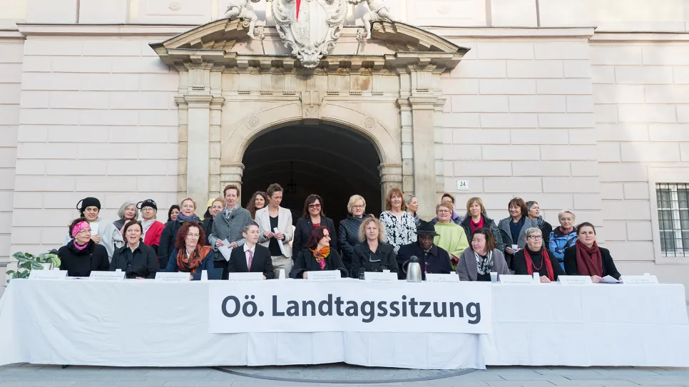 Frauen vor einem Gebäude mit Banner "Oö. Landtagssitzung", lächelnd und bunt gekleidet.