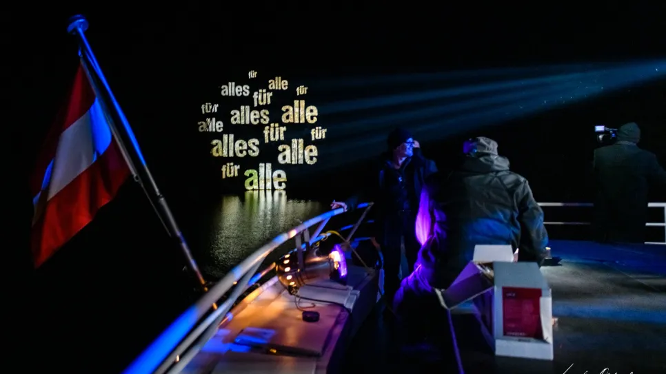 Boot bei Nacht mit Lichtprojektion "alles für alle", Menschen an Deck, österreichische Flagge.