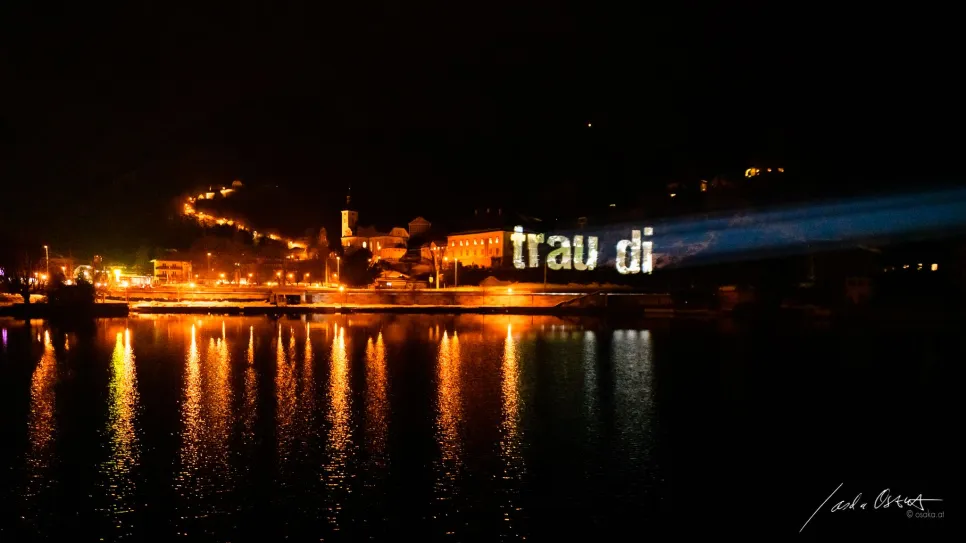 Nächtliche Stadtansicht mit beleuchtetem Schriftzug "trau di" über Wasser.
