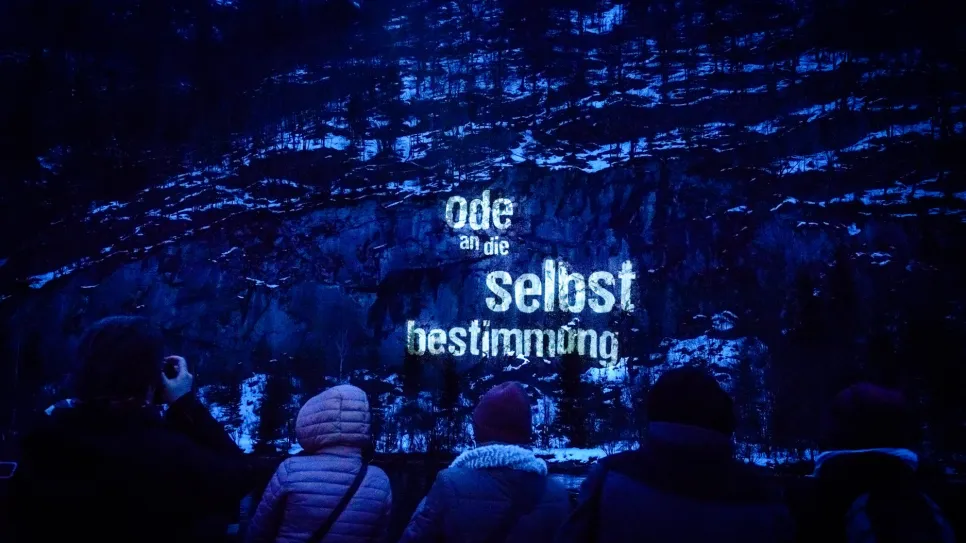Personen vor beleuchteter Felswand mit Text "Ode an die Selbstbestimmung".