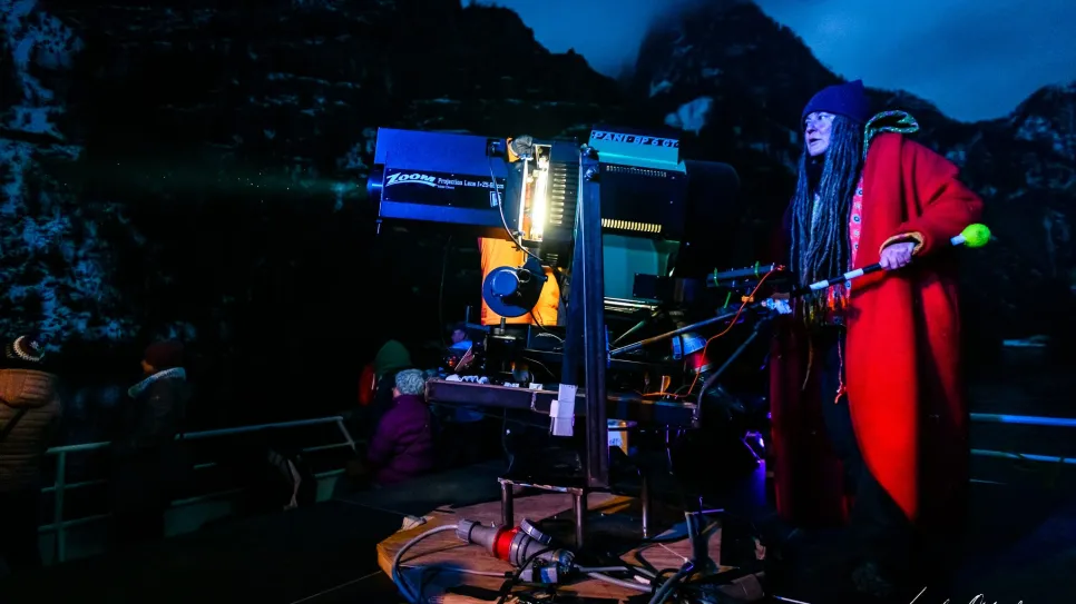 Frau mit roten Mantel bei Nacht an Filmkamera, dunkle Berglandschaft im Hintergrund.