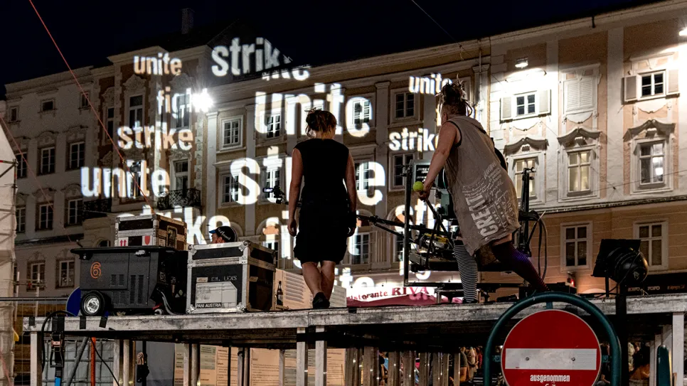 Zwei Personen auf einer Bühne, projizierte Wörter "unite" und "strike" im Hintergrund.
