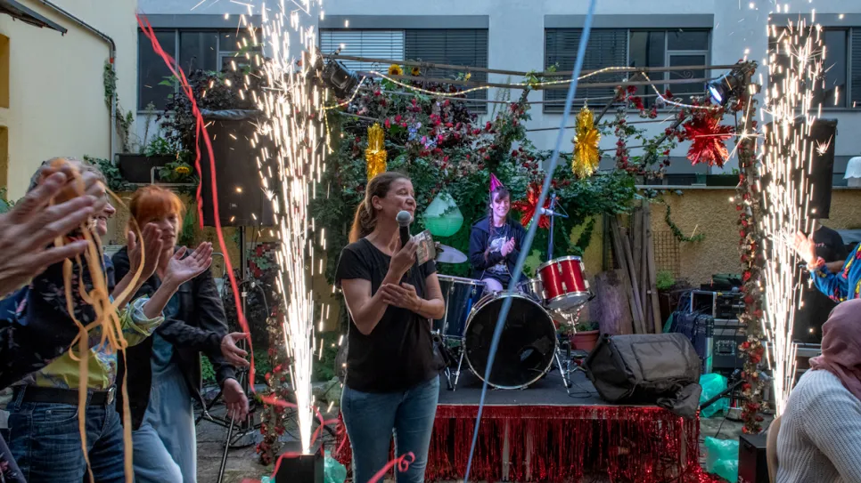 Eine Frau mit Mikrofon vor Bühne mit Band und bunten Lichtern, umgeben von Feiernden.