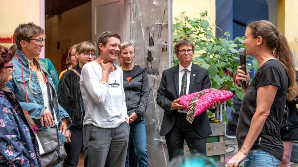 Menschen in einem Innenhof, Frau mit Mikrofon spricht zu lächelnder Gruppe.