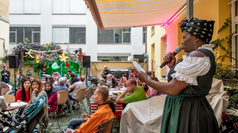Frau spricht bei Veranstaltung im Hof, buntes Publikum, festliche Dekoration.