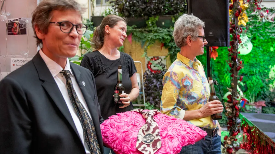 Drei Menschen bei einer Veranstaltung im Freien, mit Blumen und einem pinken Kissen.