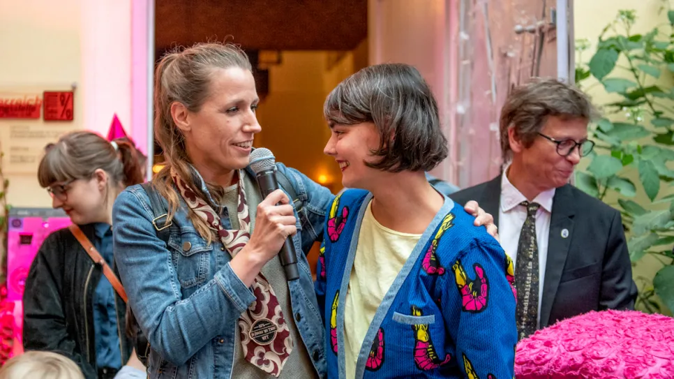 Zwei Frauen bei einer Veranstaltung, eine spricht in ein Mikrofon, umgeben von Zuschauern.