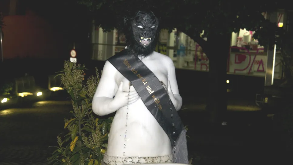 Skulptur mit Wolfskopfmaske und Schärpe, nachts beleuchtet.