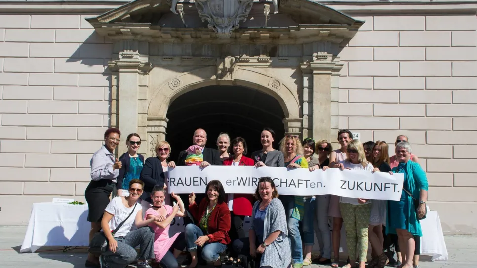 Eine Gruppe lächelnder Menschen hält ein Banner vor einem historischen Gebäude.