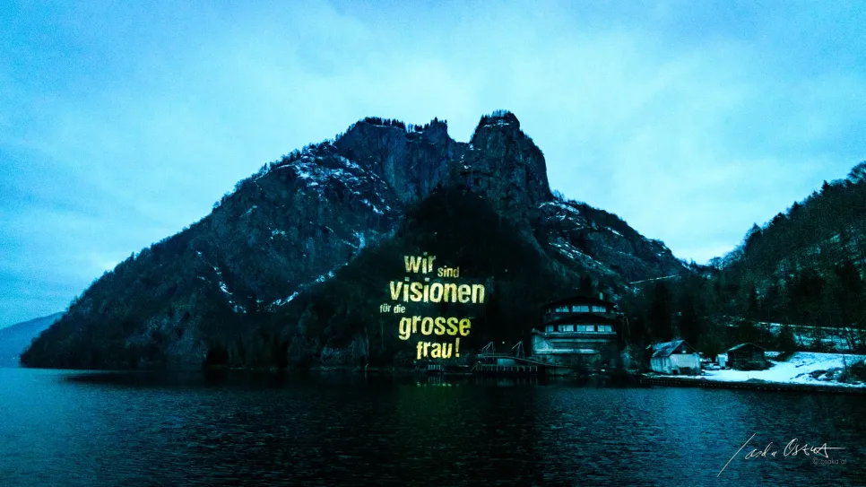 Beleuchtete Schrift auf Berglandschaft bei Dämmerung, See im Vordergrund.