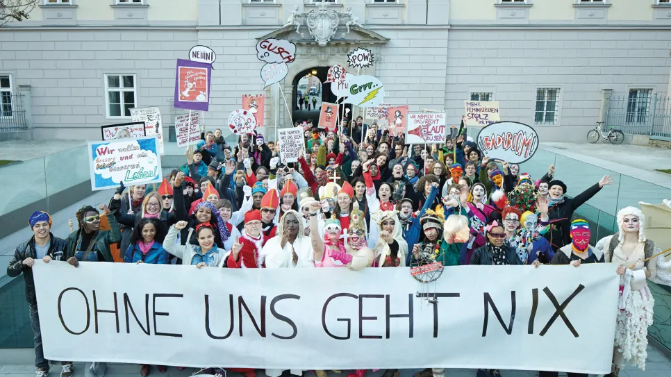 Große Gruppe Menschen mit Schildern und einem Transparent vor einem Gebäude.