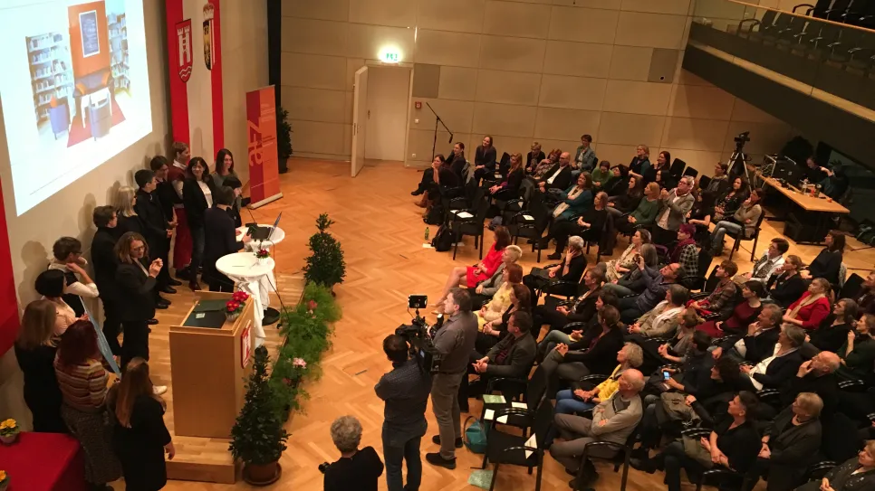 Mehrere Personen auf einer Bühne vor Publikum in einem Veranstaltungsraum.
