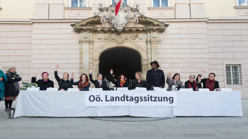 Personengruppe sitzt hinter einem langen Tisch vor einem Gebäude. Einige heben die Hand. Fotografiert.