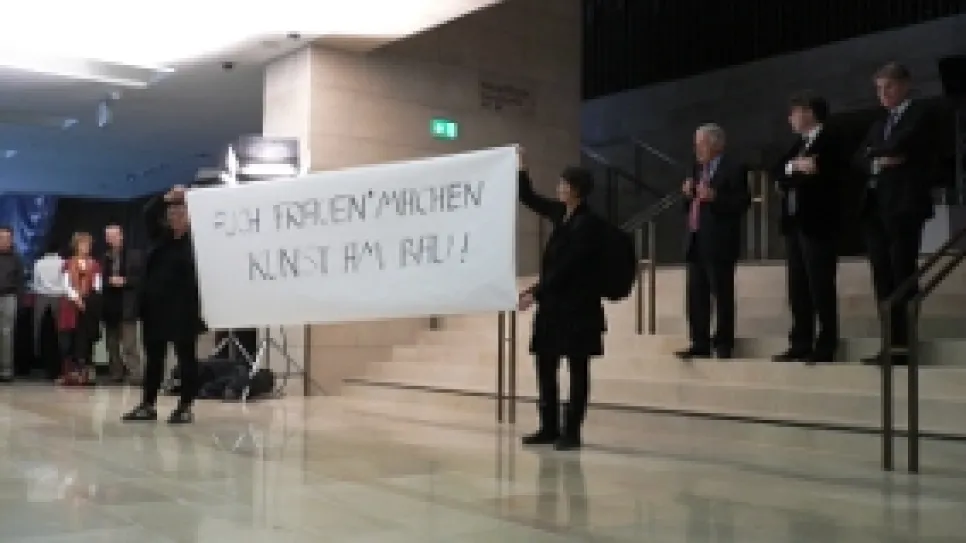Personen mit Transparent in modernem Gebäude, Publikum im Hintergrund.