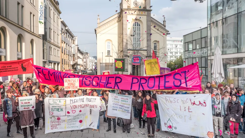 Menschen demonstrieren mit Bannern und Schildern auf einer belebten Straße vor einer Kirche.