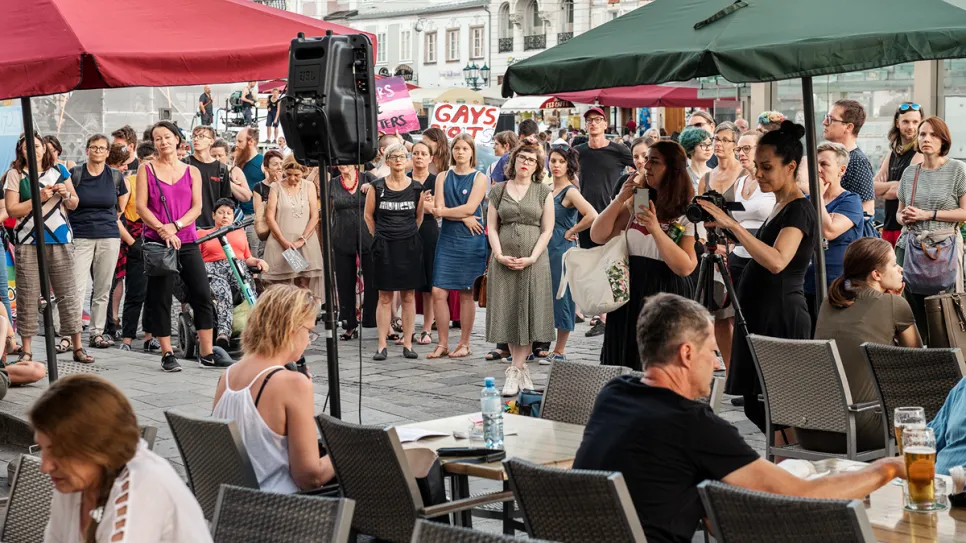 Menschenmenge auf einem Platz, einige halten Schilder, im Vordergrund Personen an Tischen.