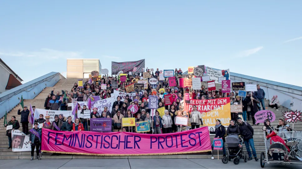 Protestierende Menschen auf Treppe mit Schildern und Banner, klarer Himmel.
